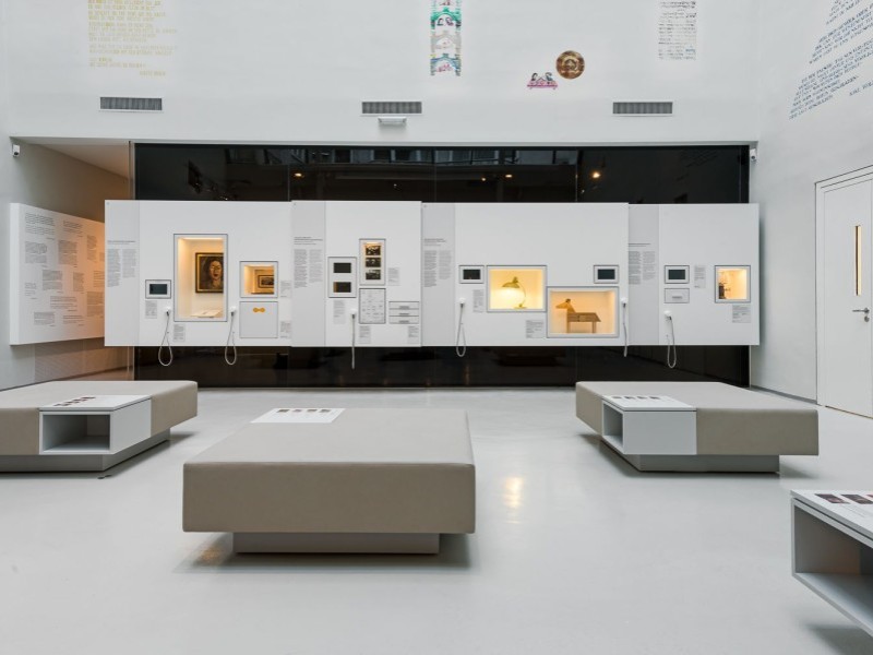 Innenraum einer Ausstellung mit weißen Wänden, eingebauten Vitrinen mit Objekten, mehreren grauen Sitzbänken auf betonfarbenem Boden