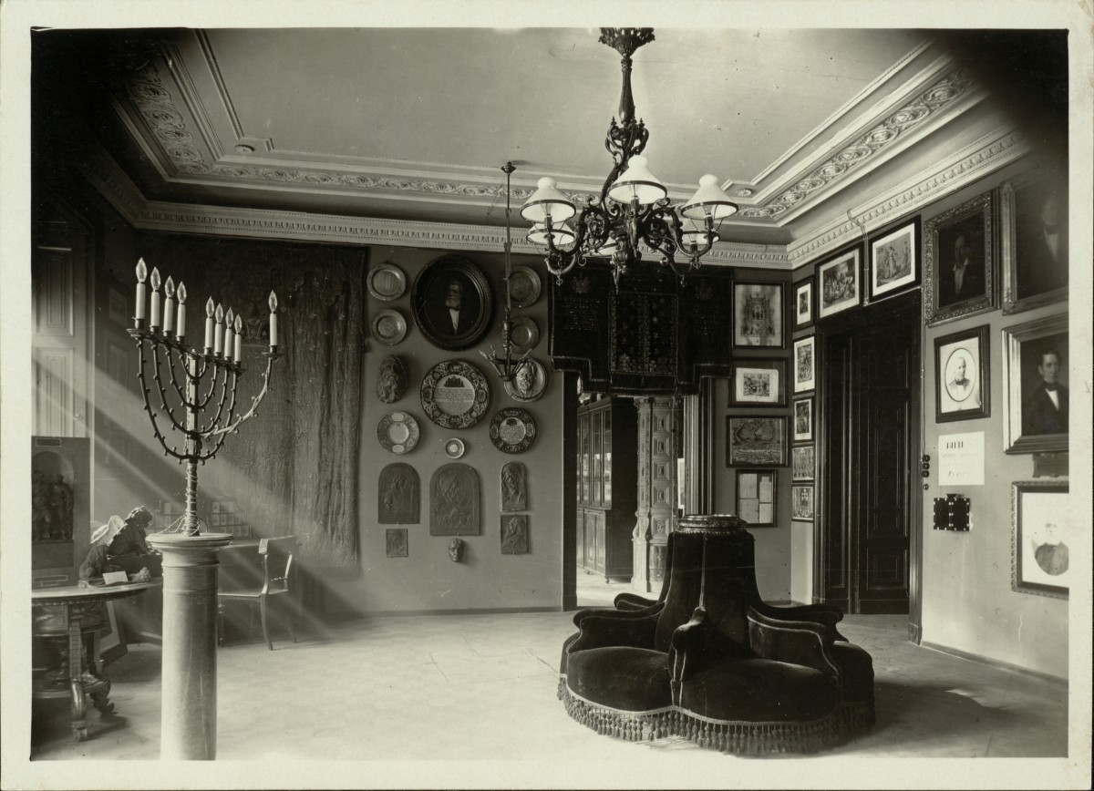 Historic museum room with menorah: Historic room with menorah on pedestal, wall plaques, numerous framed pictures, and circular seating furniture in the center
