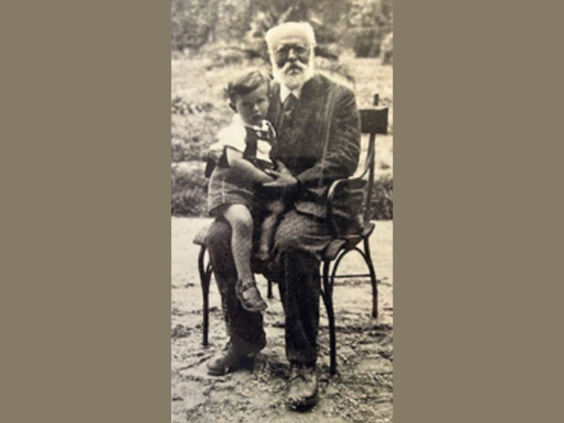 Older man with beard sitting on a chair outdoors holding a child on his lap.