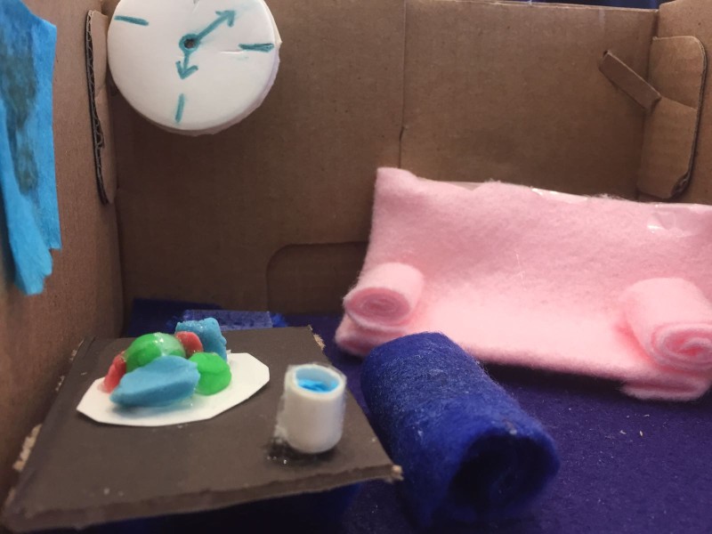 Miniaturmodell eines Wohnzimmers aus Pappe mit pinkem Sofa, blauem Teppich, Tisch mit bunter Dekoration und Uhr mit Pfeilen an der Wand