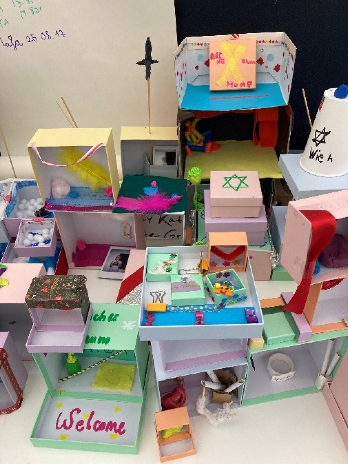 : Mehrere bunte, handgefertigte Miniaturmodelle aus Papier und Karton, darunter ein Modell mit einem grünen Davidstern und ein Modell mit der Aufschrift 'Welcome'.