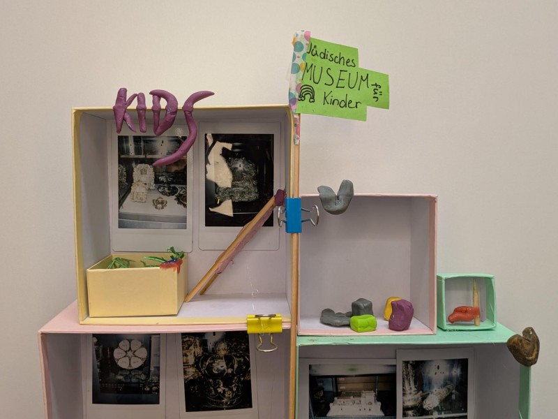 Small diorama with the word "KIDS" made of purple modeling clay, two photos in the background, and a small yellow box with green decoration in the foreground.