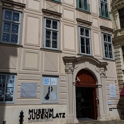 Museum Judenplatz Fassade: Fassade des Museums Judenplatz mit fünf Fenstern und dunklem Eingang unter einem ornamental verzierten Bogen, mit Schildern und Plakaten am Gebäude